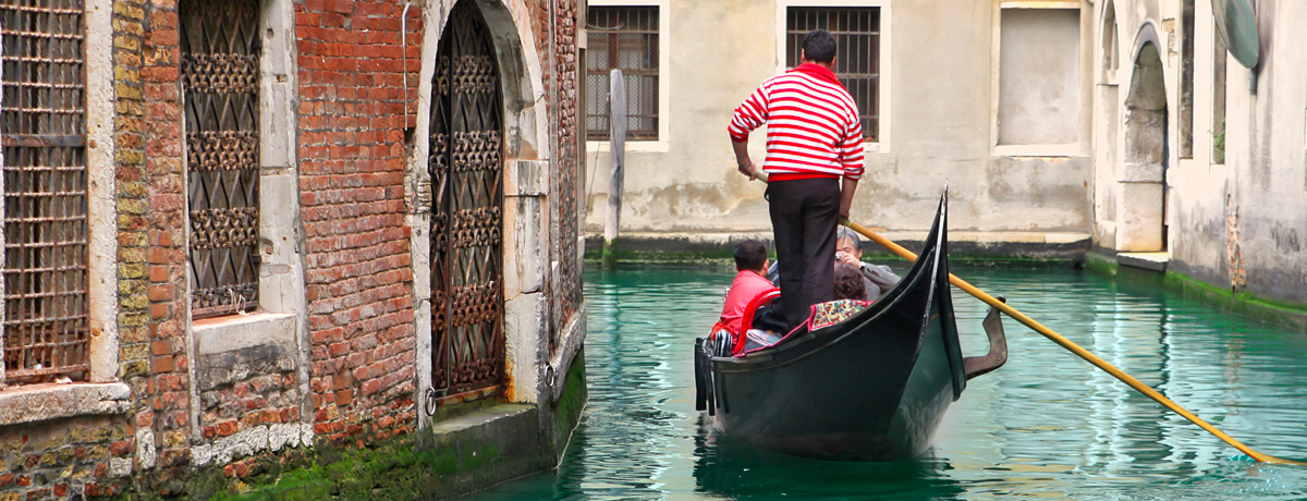 Gondola with tourists passing on narrow canal among old houses in Venice